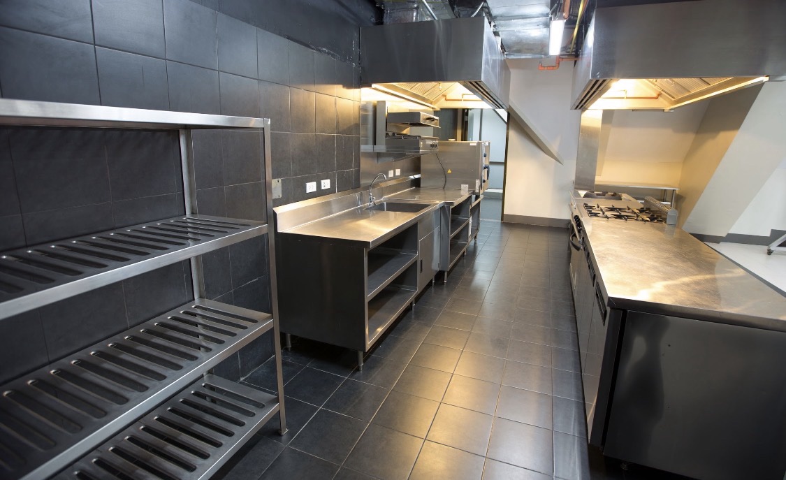 Commercial kitchen with stainless shelving, sink, hoods, and work surfaces.